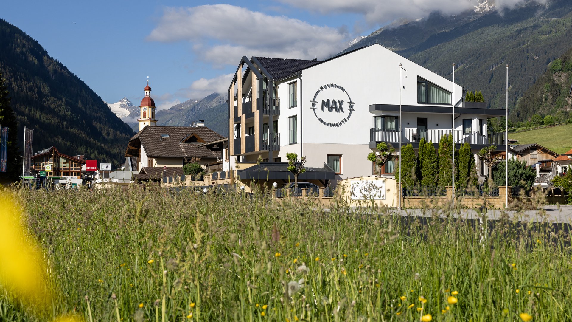 Deine Ferienwohnung in Neustift ✓ Wiese mit Blick auf moderne Alpenwohnungen und Kirche in Berglandschaft